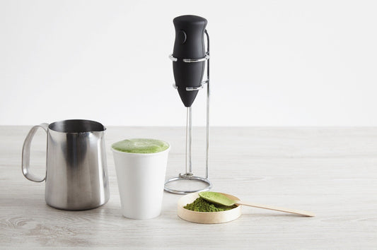 An electric milk frother stands on a table beside a matcha latte and a metal pitcher.