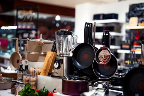 A store featuring a diverse selection of kitchen essentials, including non-stick frying pans, countertop blenders, pots, spice jars, and fresh produce.